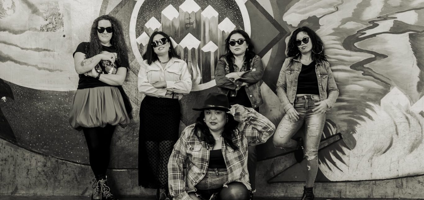 Five women posing in front of a colorful mural, wearing sunglasses and stylish outfits.